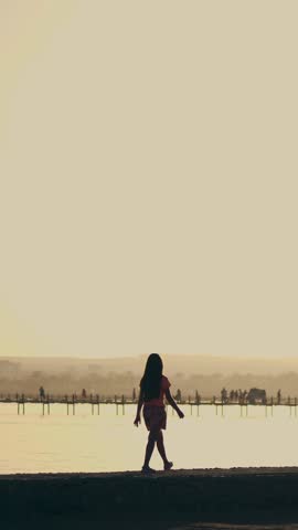 In the summer at sunset, in the bay, a teenager girl walks along the embankment. The orange sunset and the outlines of the child, palm trees on the beach