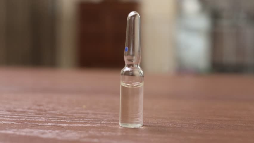 small glass ampoule filled with clear liquid standing upright on a wooden surface, with soft background bokeh and subtle rack lighting reflections dancing across the curved glass. The footage captures
