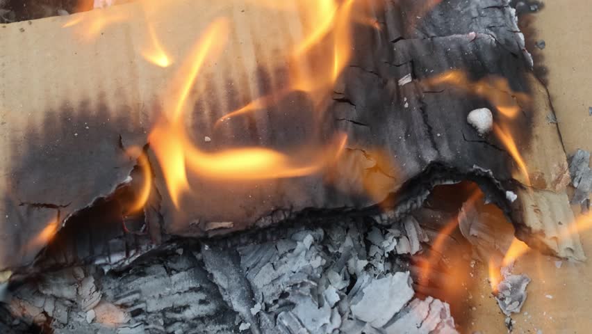 close-up of charred paper edges engulfed in flickering orange flames atop a cardboard surface, capturing the ember glow, curling ash layers, and dynamic movement of fire tongues licking the burnt 