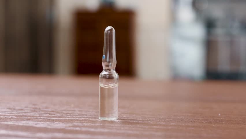 small glass ampoule filled with clear liquid standing upright on a wooden surface, with soft background bokeh and subtle rack lighting reflections dancing across the curved glass. The footage captures