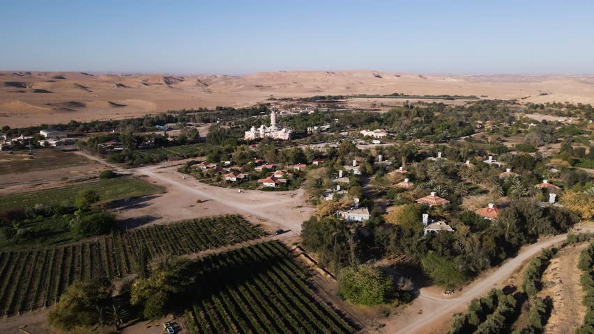 Wide view of Neot Smadar kibbutz with its iconic Art Center surrounded by agricultural fields, orchards, and desert hills under clear sky, drone slow orbiting