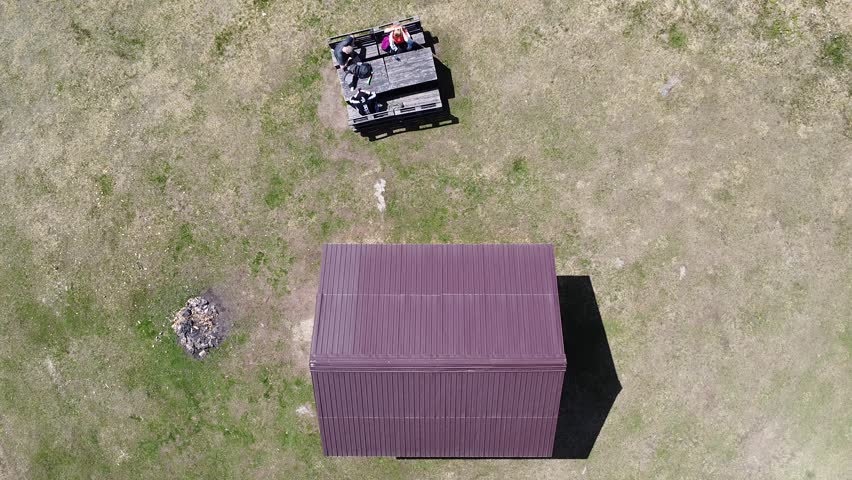 A drone view of people sitting on wooden bench near a cottage in wide countryside field on a sunny day