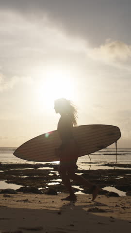 Slow Motion Silhouette Of Female Surfer Running Along Beach At Golden Hour Holding Surfboard Sunset Background Vertical Wide Shot Freedom Energy Coastal Lifestyle Dreamy Travel Sports Scene