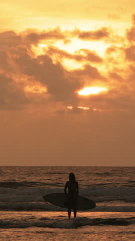 Wide Shot Silhouette Of Female Surfer Standing In Ocean Facing Horizon Orange Sunset Sky With Clouds Vertical Frame Slow Motion Peaceful Moment Surf Lifestyle Tranquil Scene Coastal Beauty