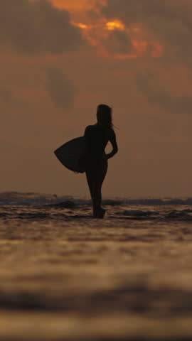 Slow Motion Silhouette Of Female Surfer Walking Out Of Ocean Holding Surfboard Windy Hair Sunset Sky In Clouds Vertical Wide Shot Coastal Mood Strength Beauty Freedom After Surf Scene