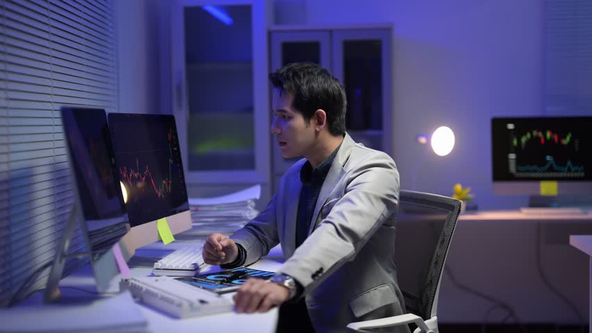A man is sitting at a desk with two computer monitors in front of him. He is wearing a gray jacket and he is working on a project
