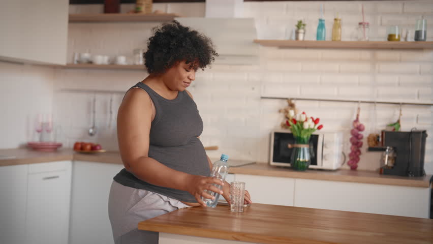 Carefree pregnant woman drinking water and using mobile phone at home kitchen. Portrait of beautiful african american woman in third trimester of pregnancy, drinking regime for good health and baby