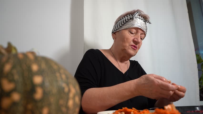 An elderly woman sorts the seeds from a fresh pumpkin. A grandmother prepares food at the table in the kitchen. Home cook