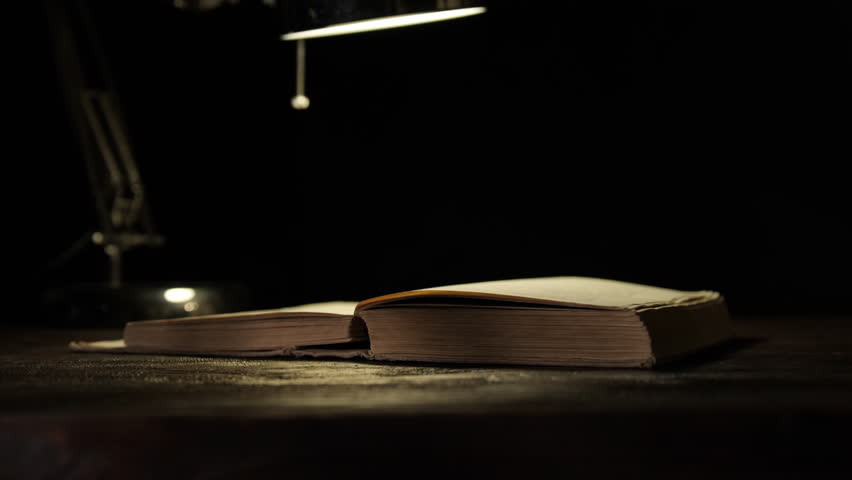 Pages of old book turning under light of table lamp on black background close-up