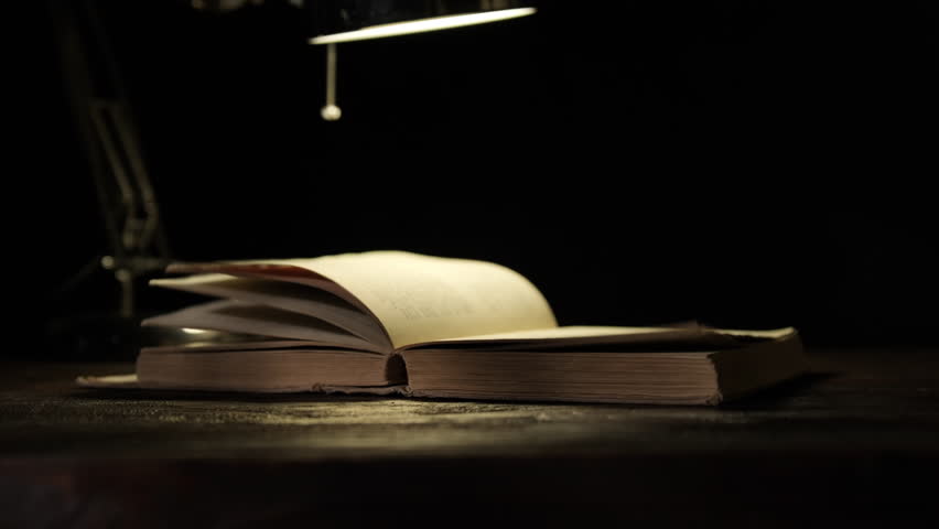 Pages of old book turning under light of table lamp on black background close-up