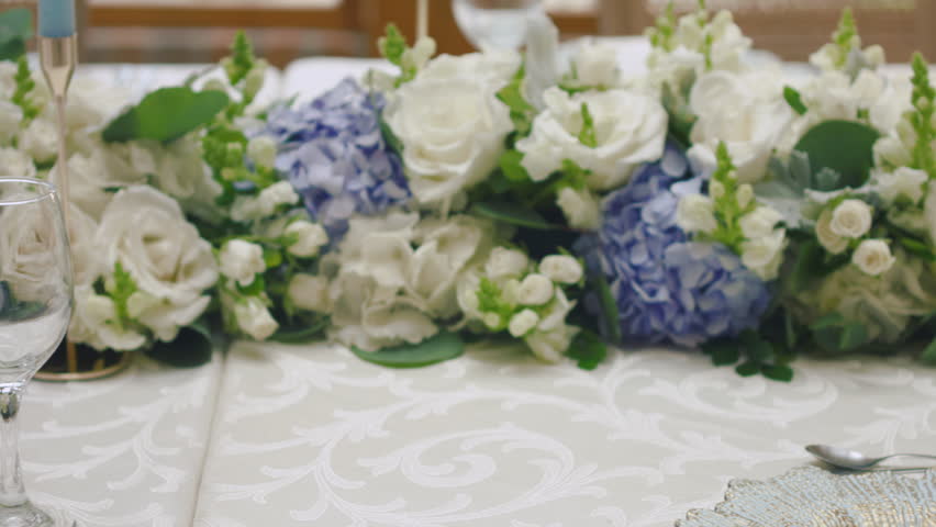 A detailed clip of an elegantly decorated wedding table. It features stemware, a white tablecloth, plates, cutlery, cloth napkins, a delicate floral arrangement, and a personalized invitation.