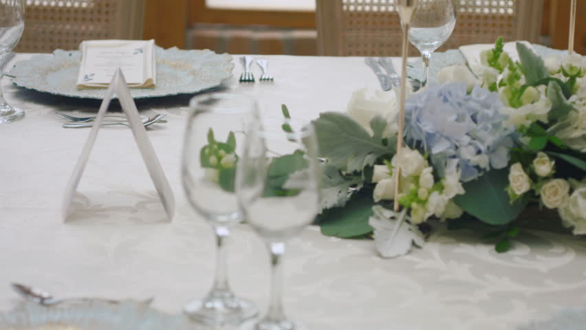 A detailed clip of an elegantly decorated wedding table. It features stemware, a white tablecloth, plates, cutlery, cloth napkins, a delicate floral arrangement, and a personalized invitation.