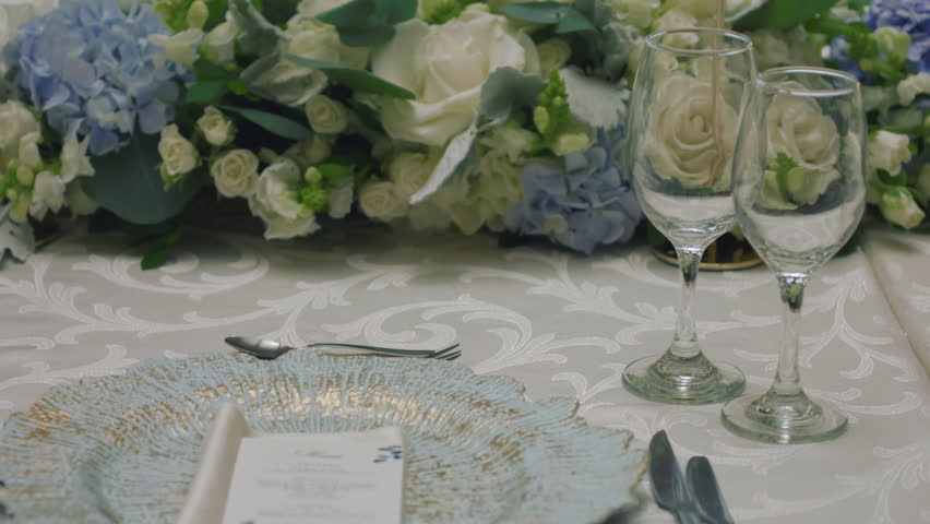 A detailed clip of an elegantly decorated wedding table. It features stemware, a white tablecloth, plates, cutlery, cloth napkins, a delicate floral arrangement, and a personalized invitation.