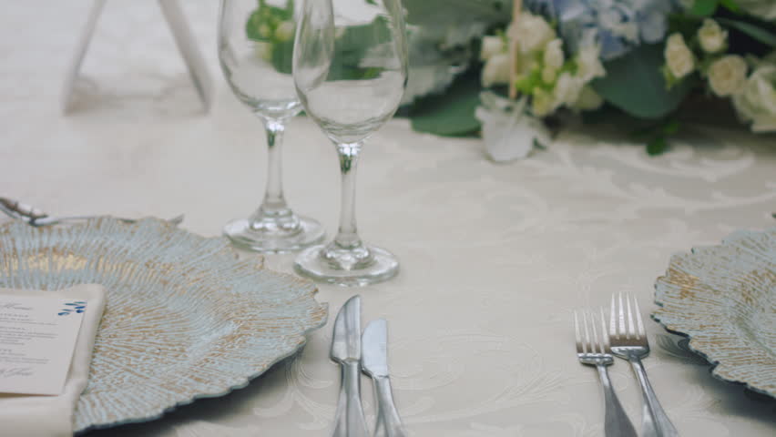 A detailed clip of an elegantly decorated wedding table. It features stemware, a white tablecloth, plates, cutlery, cloth napkins, a delicate floral arrangement, and a personalized invitation.