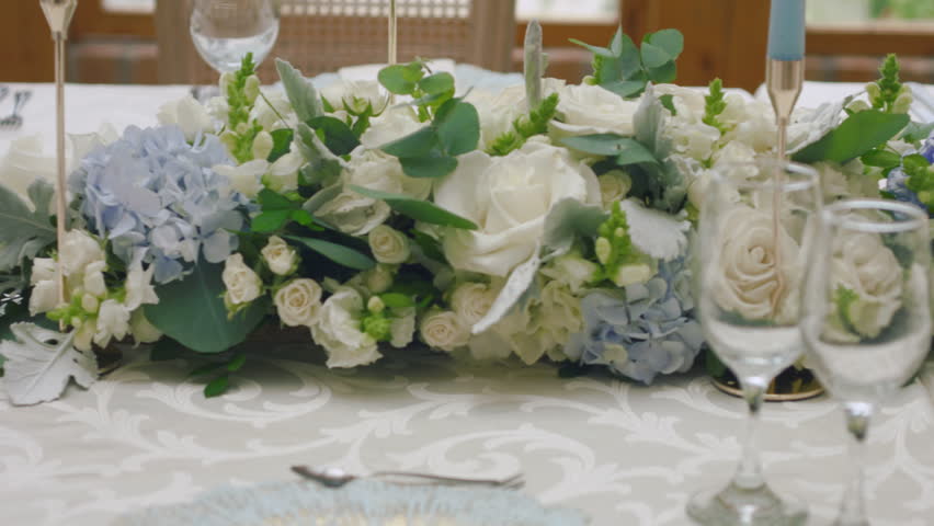 A detailed clip of an elegantly decorated wedding table. It features stemware, a white tablecloth, plates, cutlery, cloth napkins, a delicate floral arrangement, and a personalized invitation.