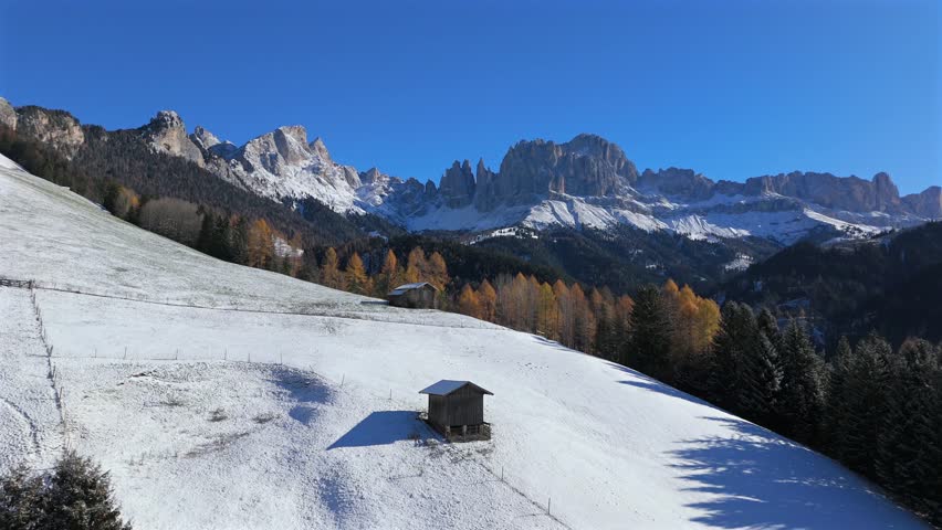 Drone footage of a snowy hillside near Tiers Tires in South Tyrol Alto Adige Italy with wooden alpine huts golden trees and the Rosengarten Catinaccio Dolomites in clear winter light