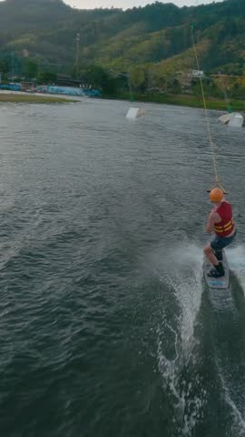 Vertical video. Engineer cable wakeboarder man in orange construction helmet trying cable wakeboard in lake park water sport. Extreme activity guy training water sport fpv drone follow aerial shot 4k