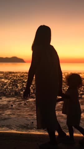 Mother and daughter walking hand in hand at sunset on the beach. Silhouetted against golden light, evoking warmth, love, and peaceful connection. Vertical format.