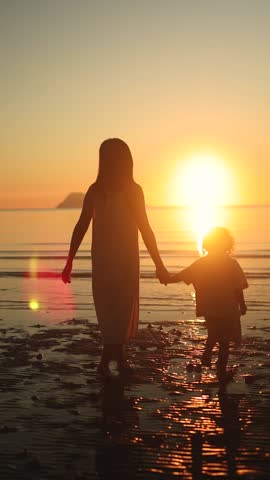 Mother and daughter walking hand in hand at sunset on the beach. Silhouetted against golden light, evoking warmth, love, and peaceful connection. Vertical format.