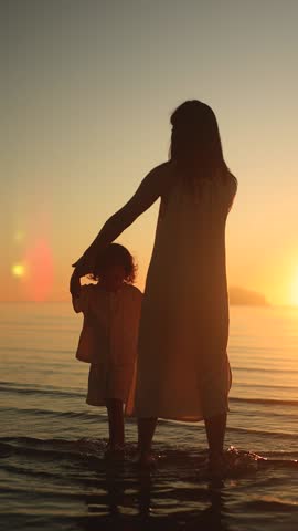Mother and daughter dancing in the sea at sunset, captured in a soft, emotional silhouette during golden hour. Heartfelt and cinematic moment. Vertical video.