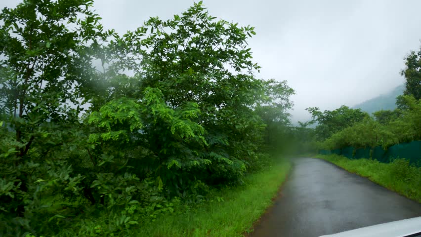 Car POV view of road with green trees and cloudy sky in rainy weather at Maharashtra, India. Raindrop on lens. Road trip through Sahyadri hills. Back to the road. 