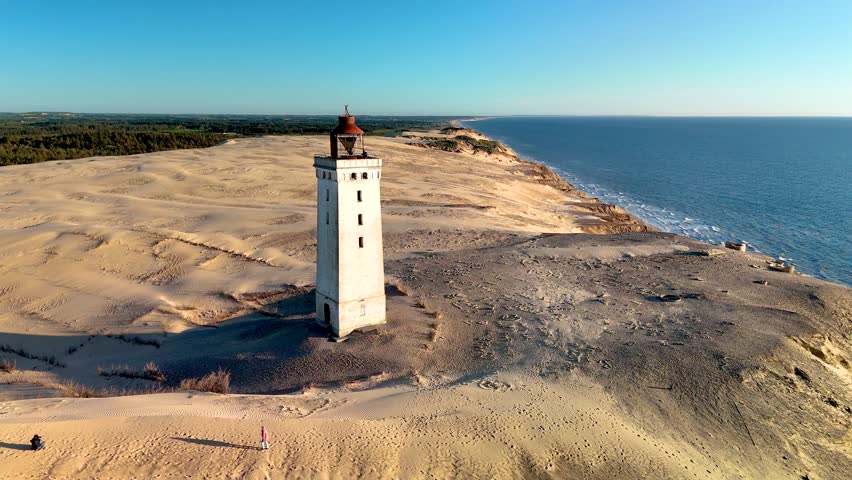 Wander through the stunning dunes of Rubjerg Knude Fyr Lighthouse at sunset in Jutland, Denmark. The serene landscape features a historic lighthouse overlooking the North Sea