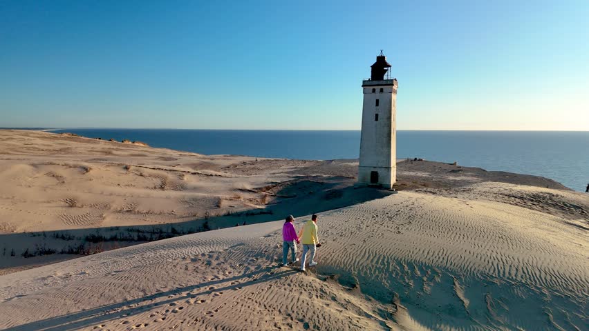 Stroll along the picturesque sands of Rubjerg Knude Fyr Lighthouse in Jutland, Denmark, as a couple enjoys the warm sunset glow near a historic lighthouse overlooking the tranquil sea, creating a