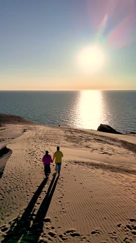 A tranquil sunset casts a warm glow over Rubjerg Knude Fyr Lighthouse in Jutland, Denmark. A couple holds hands as they walk along the secluded sandy shore, enjoying the peaceful ocean waves.