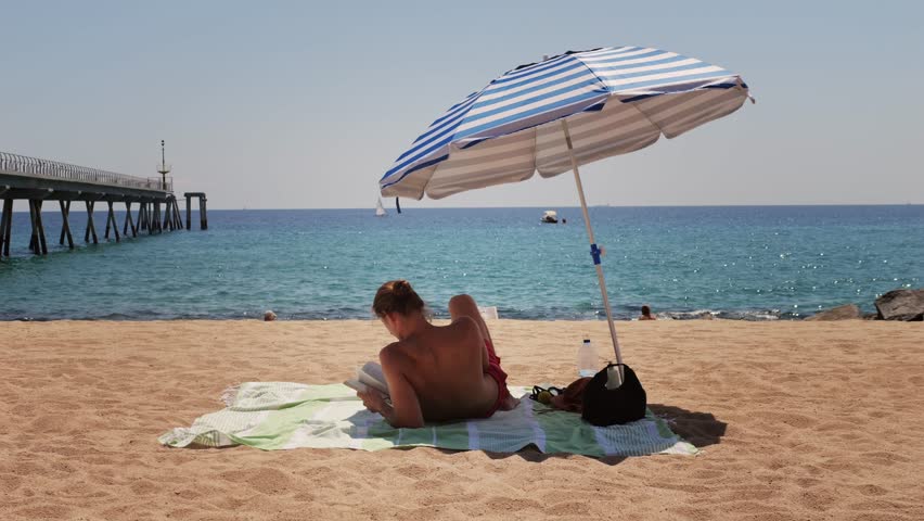 Man Tourist Enjoying a Relaxing Day at the Beach, Comfortably Seated Under a Bright Umbrella, Barcelona, spain.