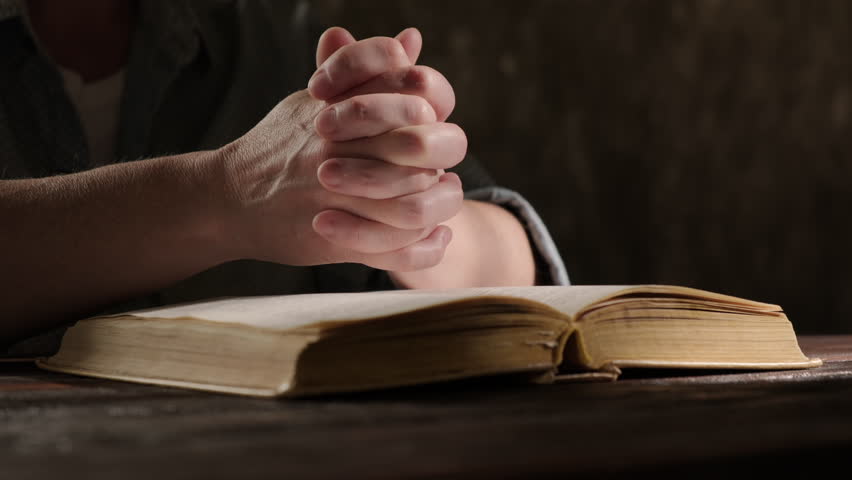 Clasped hands in prayer over open book on table close-up