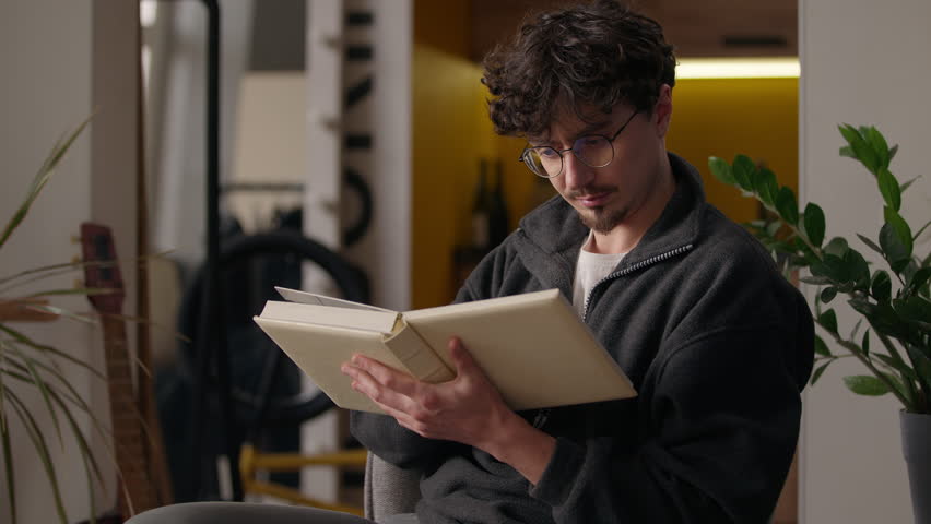 Focused Caucasian guy reading book at home hipster male read story literature paper knowledge reader in glasses studying information library hobby man learn homeowner holding textbook notebook leisure