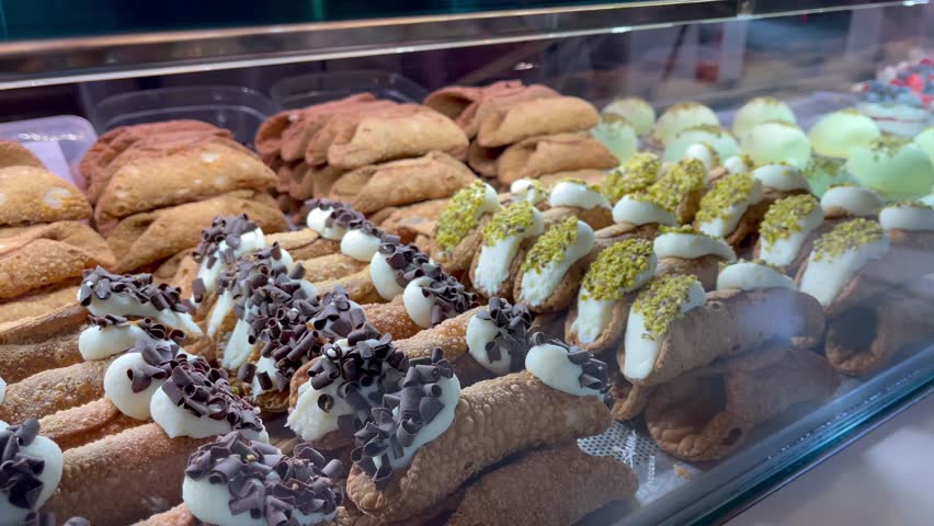 Assorted traditional Sicilian cannoli pastries filled with ricotta cream and decorated with chocolate chips, pistachios, and candied orange, displayed in a bakery glass case
