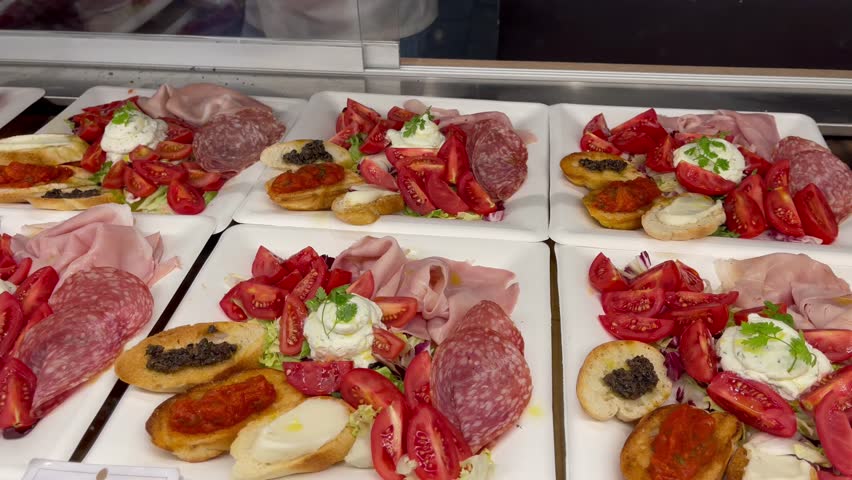 Assorted Italian antipasto plates with cold cuts, cherry tomatoes, crostini, and cheese on white platters, displayed at a deli counter or Italian restaurant