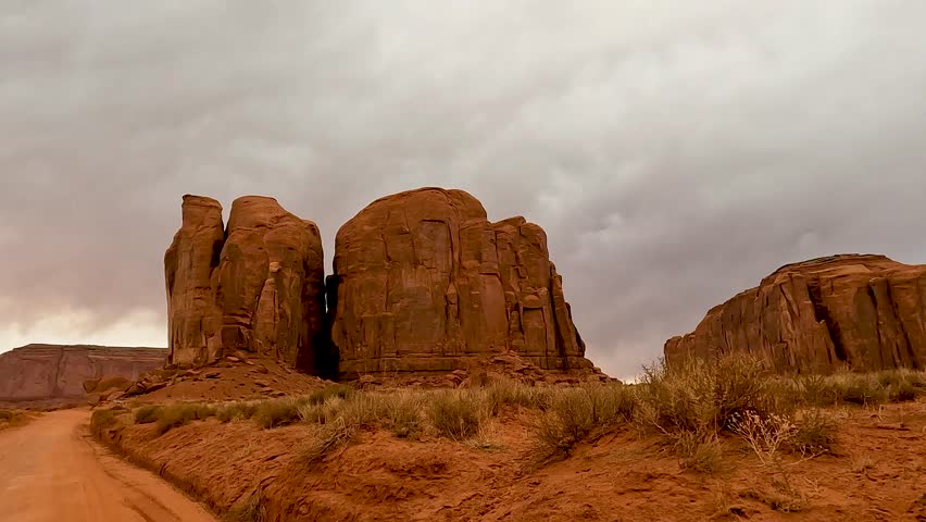 Cinematic POV shot of a car driving along a scenic desert road in Monument Valley, heading towards the Thumbs and other iconic sandstone formations. The vast red landscape, towering buttes, and expans