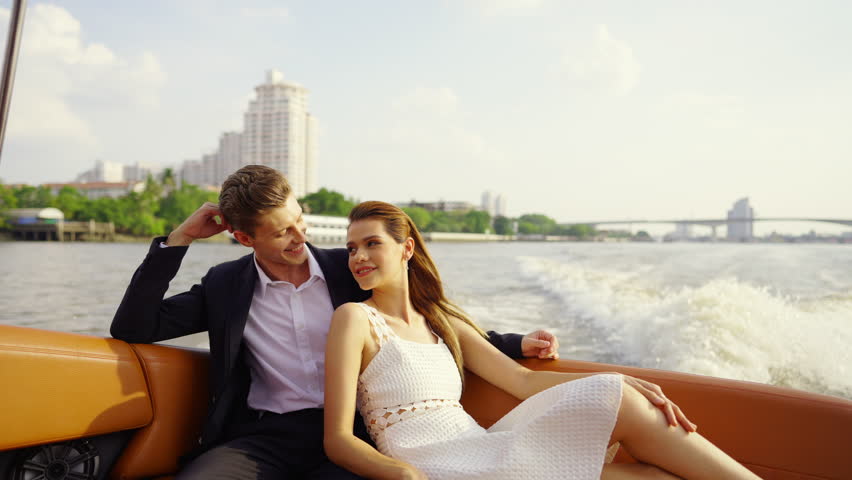 Happy Caucasian man and woman couple enjoy urban lifestyle travel city on luxury private boat yacht sailing in the river and looking beautiful cityscape skyline at sunset on summer holiday vacation.