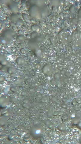 Vertical footage, View from below of tumultuous current of air bubbles sparkling under the water surface against the background of the sun and the blue sky, backlight macro video, slow motion