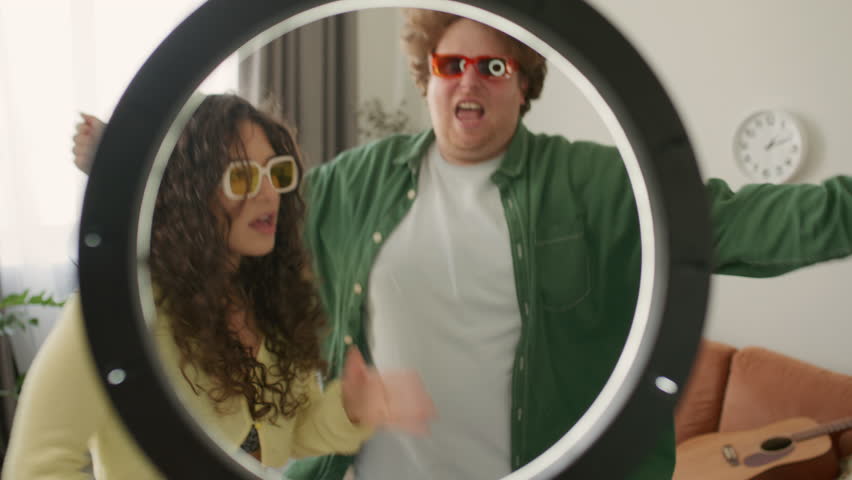 Young creative man and woman wearing colorful sunglasses, dancing excitedly in front of ring light in living room, filming funny video content for social media. Zoom shot