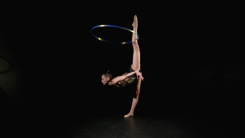 Full length of professional flexible rhythmic gymnast in stylish leotard practicing rhythmic gymnastic elements, stretching in standing side split with hoop rotating around leg on black background.