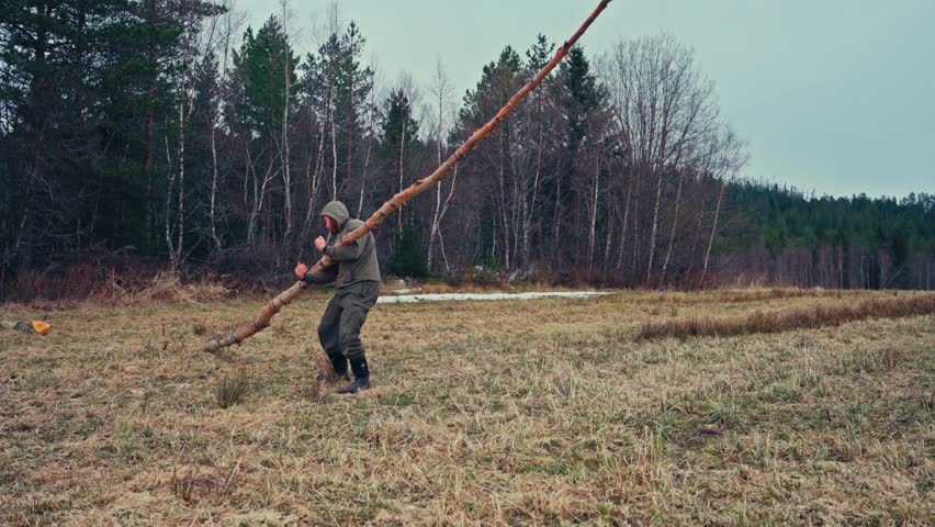 A Man is Lifting a Heavy Tree Trunk in an Open Field, Bordered by a Dense Forest - Static Shot