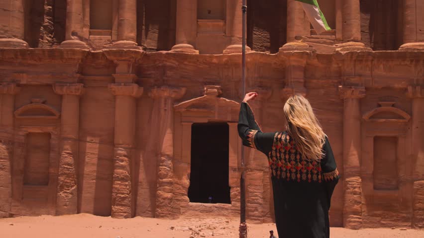 A woman stands by a flagpole with the Jordan flag, facing the ancient Treasury building in Petra, Jordan.