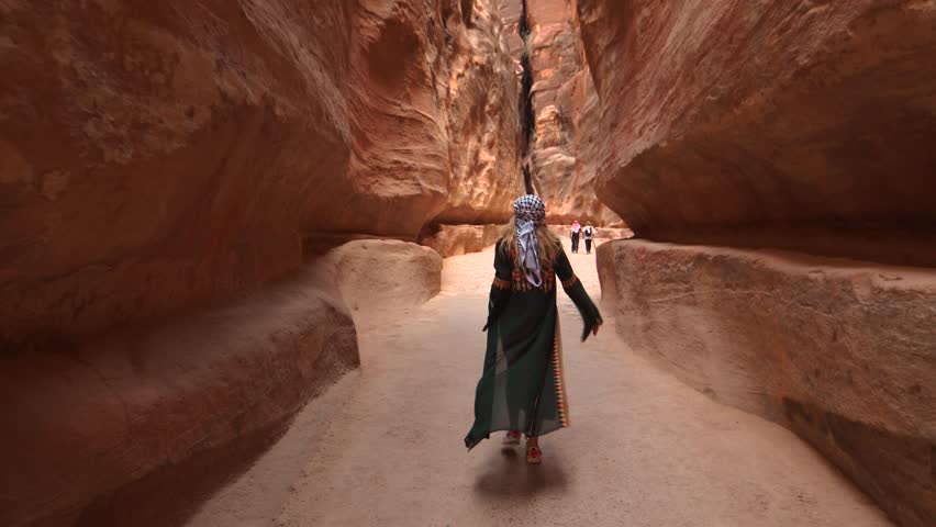 Person walking through a narrow desert canyon with towering rock walls on a sunny day, likely the Siq in Petra, Jordan.