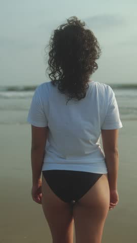 Woman in white t-shirt and black bikini bottoms stands facing ocean waves on sandy beach