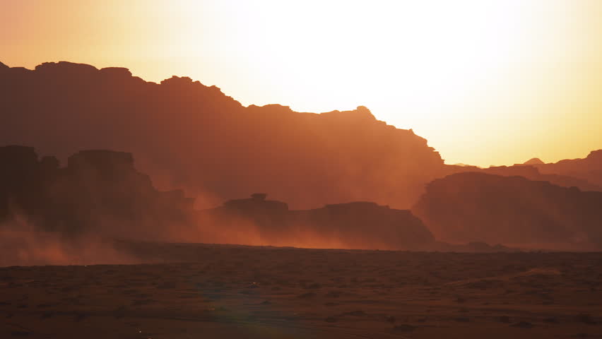Desert landscape with rock formations silhouetted against the sky at sunrise or sunset with dust blowing across the ground in Wadi Rum, Jordan.