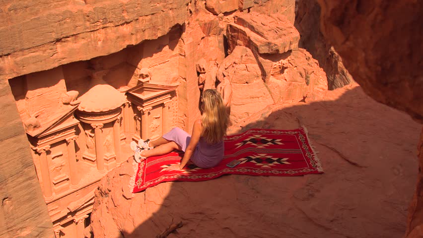 Woman sits on a red rug overlooking Al-Khazneh at the ancient city of Petra, Jordan.