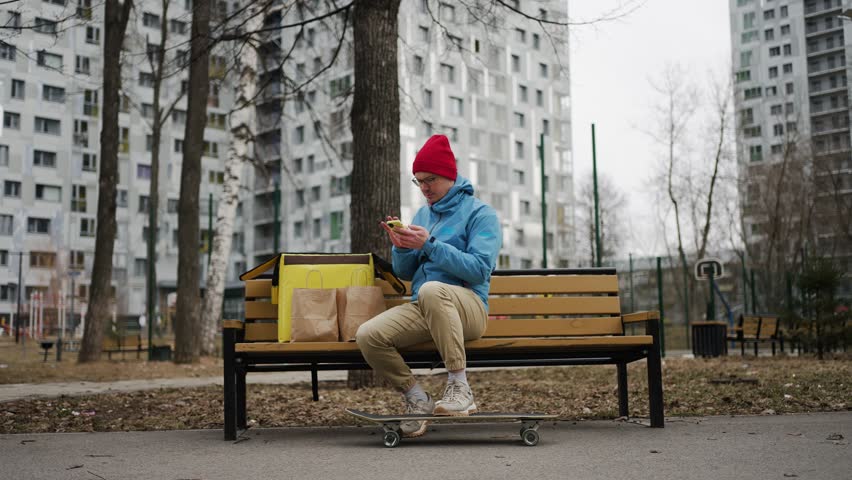 Food delivery professional resting on skateboard in urban park, preparing thermal bag and checking orders via smartphone while waiting for next delivery assignment
