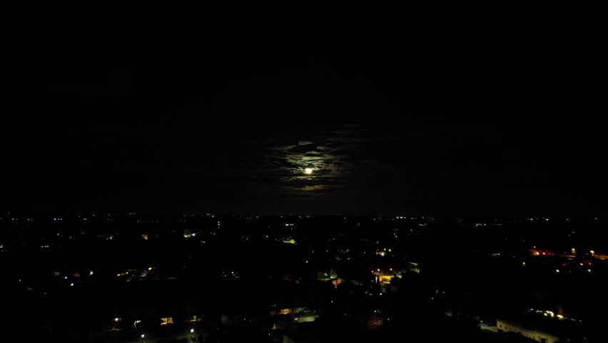 A rising-up drone footage of the cityscape illuminated at night, with the full moon glowing between the clouds in the dark sky in background