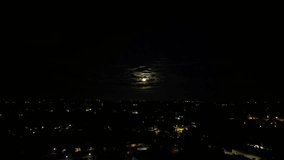 A rising-up drone footage of the cityscape illuminated at night, with the full moon glowing between the clouds in the dark sky in background - Powered by Shutterstock - Get 15% off with code: PIKWIZARD15