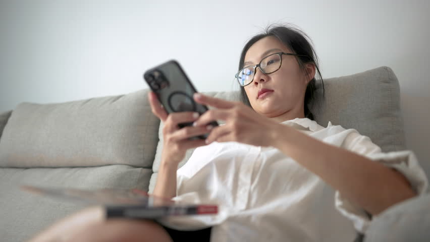 Tired Asian woman in white shirt rubs her eyes while resting on couch after screen time. A realistic and emotional moment of fatigue and digital burnout in modern lifestyle.