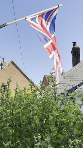 A United Kingdom, UK, Union Jack flag, in red, white and blue, hanging outside from a pole, and blowing in the wind between two houses in a suburban area.