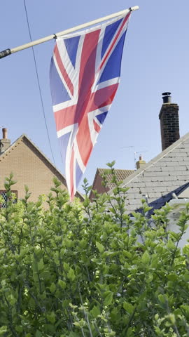 A United Kingdom, UK, Union Jack flag, in red, white and blue, hanging outside from a pole, and blowing in the wind between two houses in a suburban area.